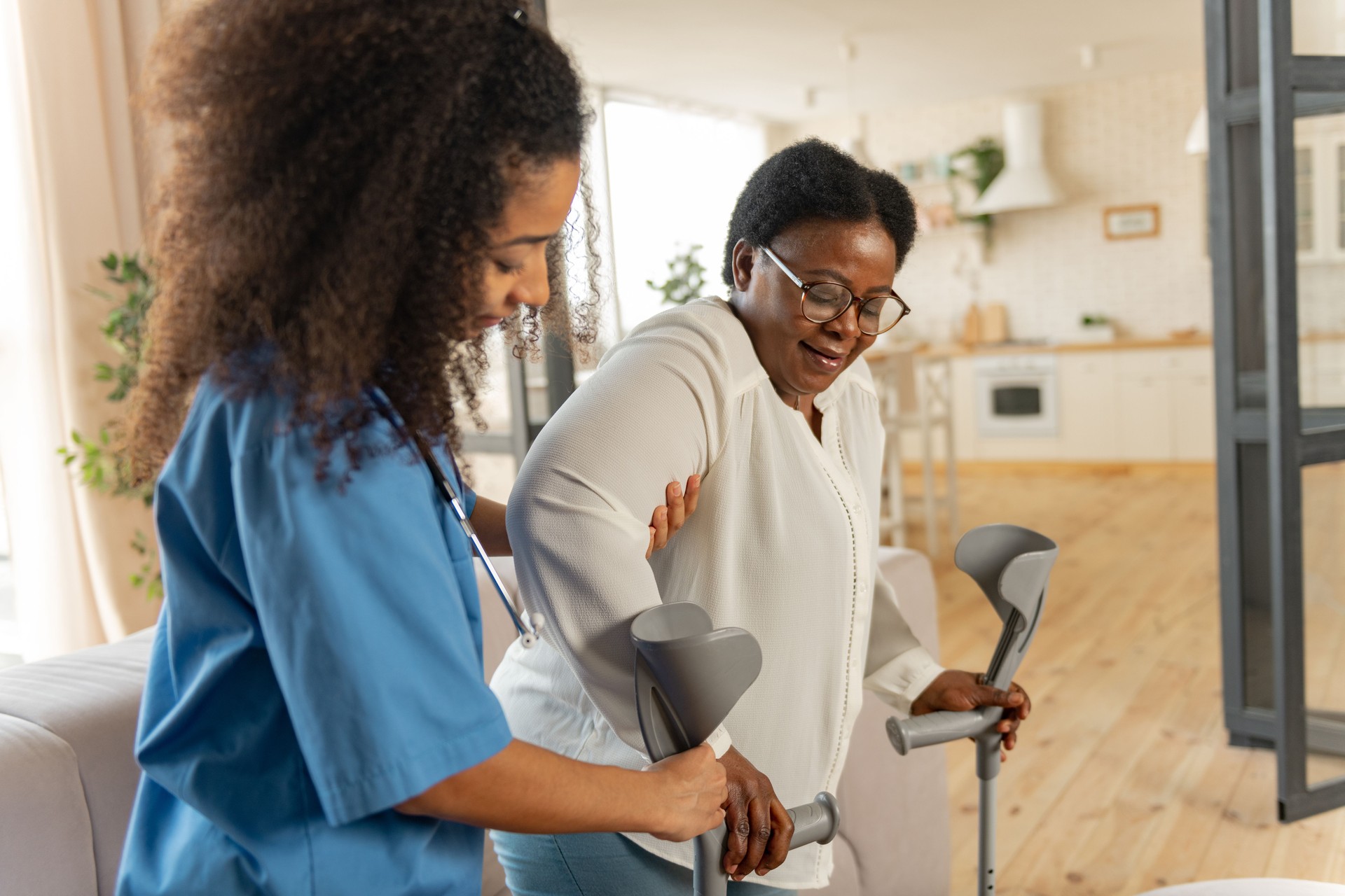 Curly young nurse giving crutches to aged woman after leg surgery