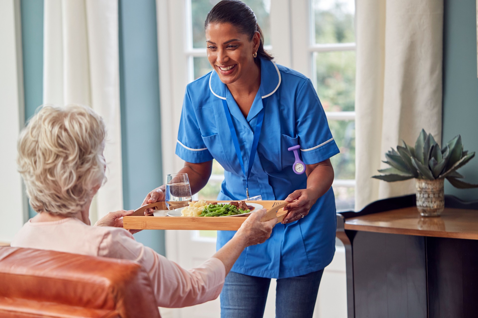 Female Care Worker In Uniform Bringing Meal On Tray To Senior Woman Sitting In Lounge At Home